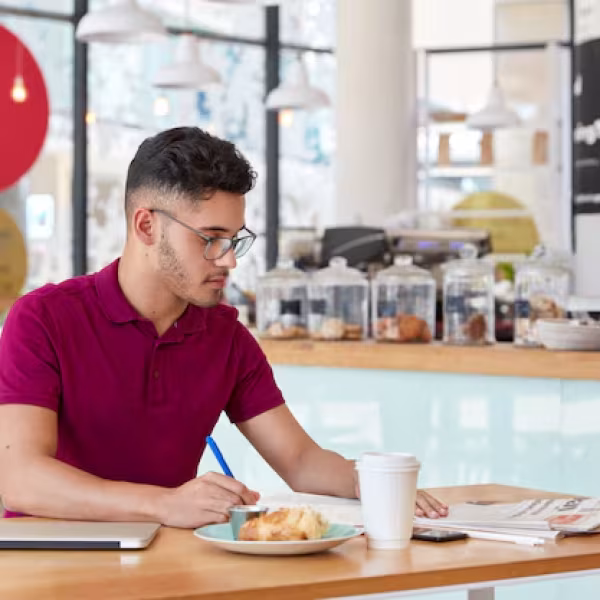 photo-stylish-man-with-trendy-haircut-writes-records-notepad-focused-newspaper-drinks-takeaway-coffee-uses-modern-laptop-computer-freelance-work-hipster-guy-makes-recordings_273609-29250